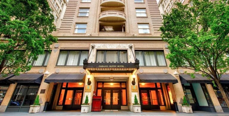 The exterior of Embassy Suites Hotel, featuring a grand entrance with bright lights, tall windows, black awnings, and decorative columns, framed by green trees on both sides.