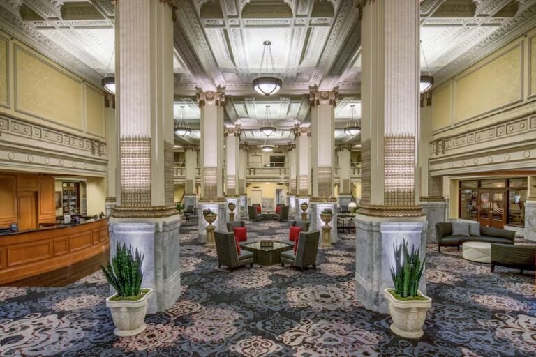 A spacious, ornate hotel lobby with tall columns, intricate ceiling details, elegant lighting, patterned carpet, seating areas with armchairs and tables, potted plants, and a wooden reception desk on the left.