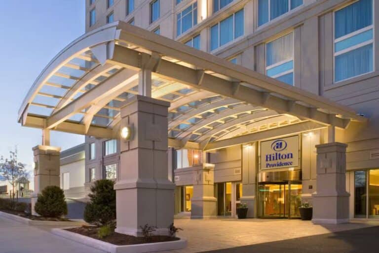 The entrance of a modern Hilton hotel with a large glass and metal canopy, illuminated by warm lights, and the Hilton Providence sign above the doors. The building has large windows and well-kept landscaping.