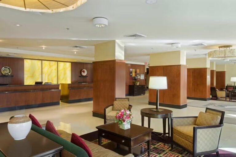 A spacious hotel lobby with wood-paneled walls, a front desk area, comfortable armchairs, a table with a flower vase, modern lighting, and decorative accents. The atmosphere is clean and welcoming.