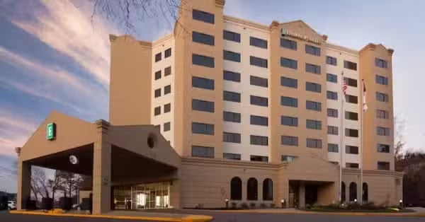 A multi-story hotel building with beige and tan exterior, numerous windows, an entrance canopy, and flags at the front, photographed under a partly cloudy sky at dusk.