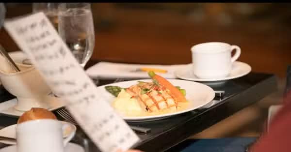 A person holds a handwritten menu in the foreground, with a plated meal of grilled fish, mashed potatoes, carrots, and asparagus, along with a cup and glass of water, visible in the background.