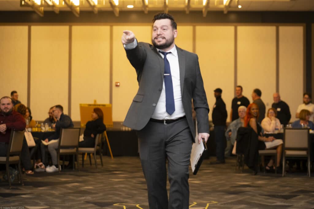 A detective character in a suit dramatically points during a live Dinner Detective interactive murder mystery performance in front of engaged guests