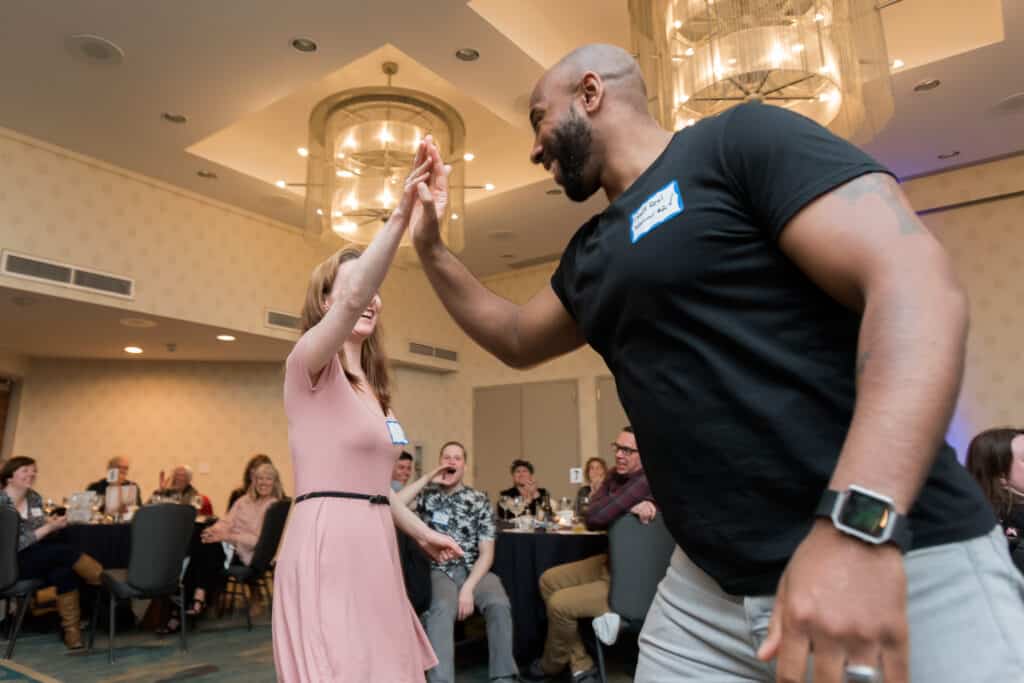 Two guests high-fiving and laughing during a live Dinner Detective murder mystery show, surrounded by engaged audience members