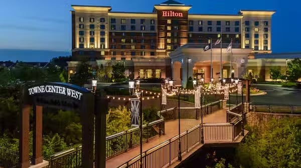 A large, well-lit Hilton hotel stands behind a pedestrian bridge decorated with string lights and Towne Center West signage, surrounded by greenery, in the evening.