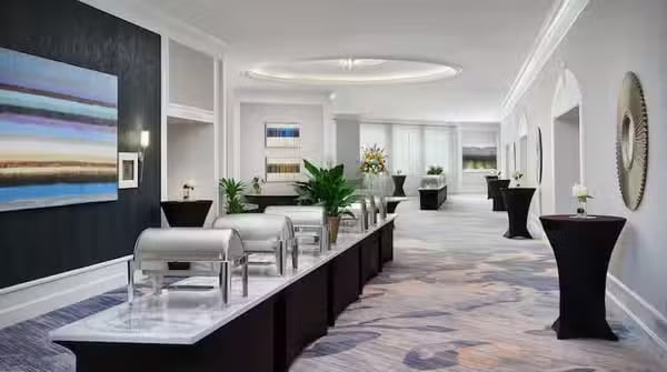 A bright, modern hallway set up for an event, featuring serving tables with silver chafing dishes, black cocktail tables, abstract art on the walls, plants, and decorative lighting.