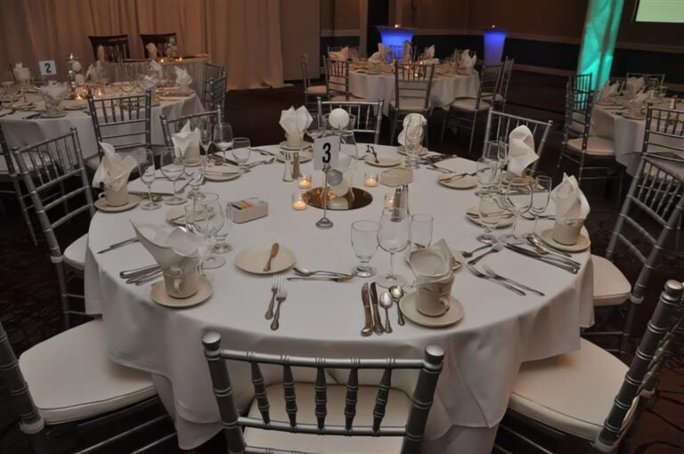 A round table set for a formal event with white tablecloth, folded napkins, cutlery, glassware, plates, and candles. Number 3 is displayed at the center. Similar tables are arranged in the background.