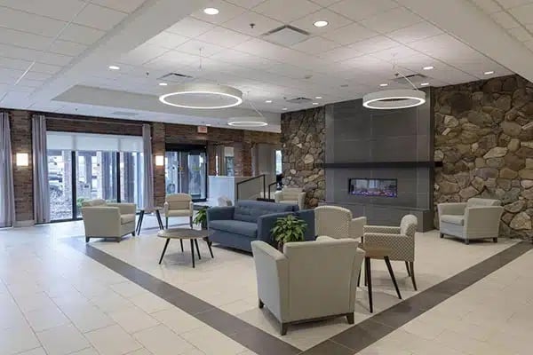 Modern hotel lobby with gray armchairs, sofas, small tables, and green plants. Large stone fireplace and circular ceiling lights add a cozy touch. Floor-to-ceiling windows provide natural light.