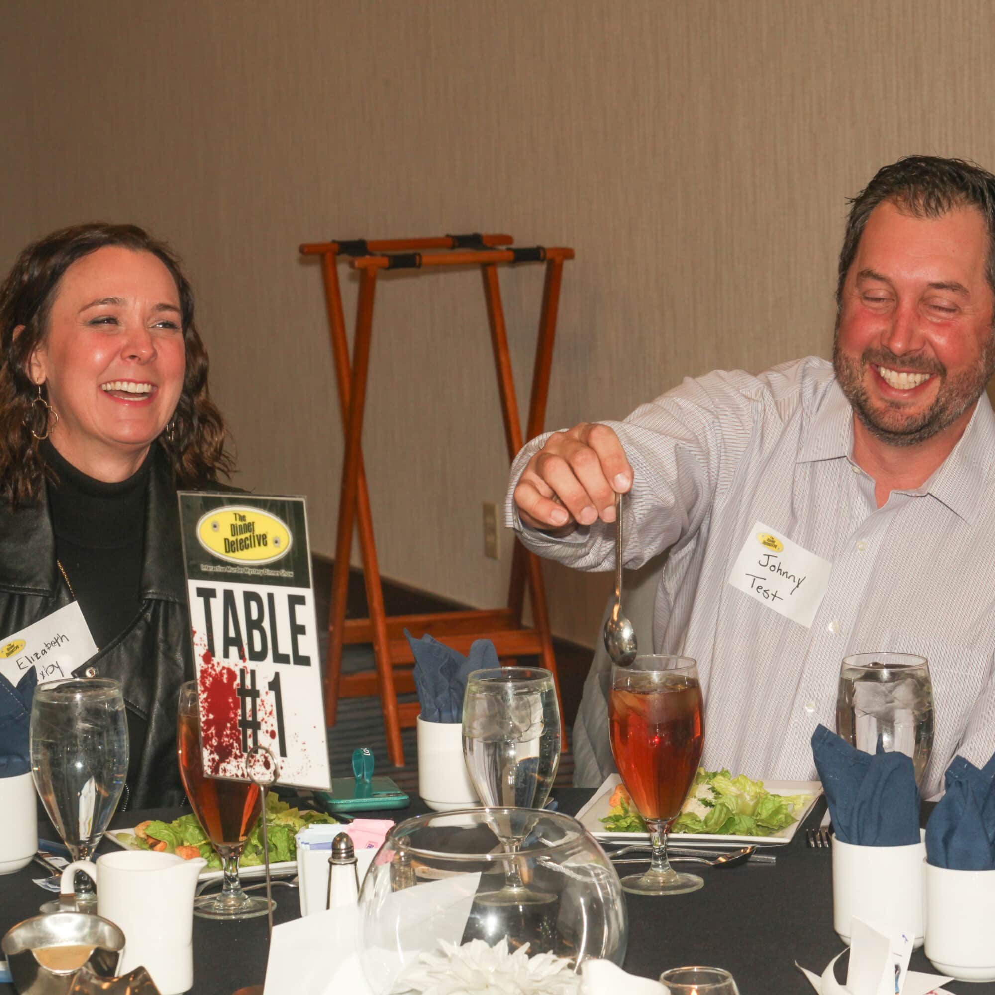 Two smiling guests enjoying dinner at The Dinner Detective Salt Lake City, seated at Table #1 with iced tea and salads.