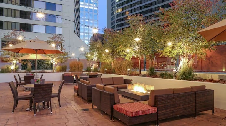 Outdoor patio area with wicker sofas and a fire pit, surrounded by potted plants and small trees. String lights are hung overhead, and nearby tall buildings are visible in the evening light.