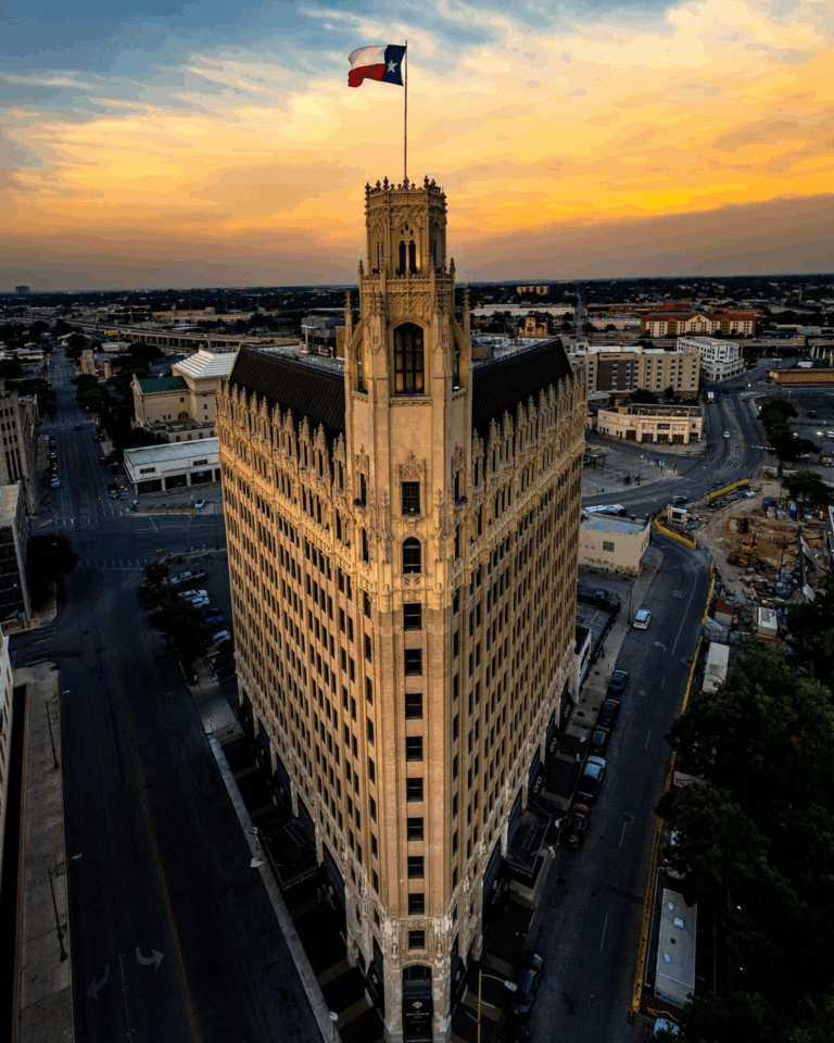 A tall, historic building with a Texas flag on top stands at a street intersection at sunset, surrounded by city streets and buildings under a colorful sky.