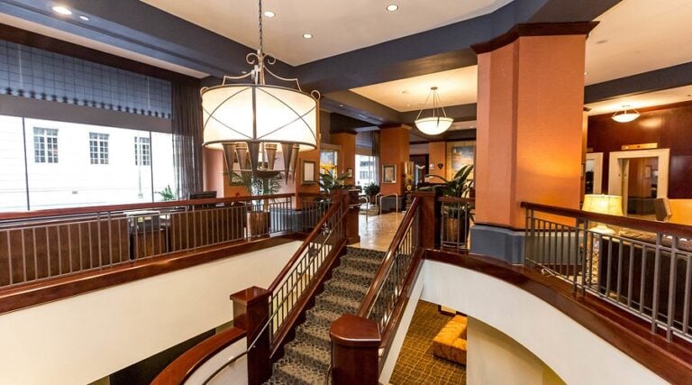 A spacious hotel lobby features a grand staircase with dark wood railings, modern chandeliers, seating areas, large windows, and warm lighting, creating an inviting and elegant atmosphere.