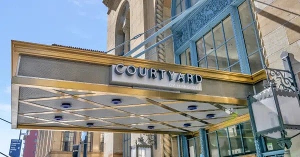 A gold and silver hotel canopy with the sign Courtyard by Marriott extends from a historic building with ornate blue metalwork and large windows.