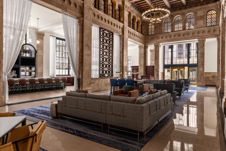 Spacious hotel lobby with tall ceilings, large windows, elegant chandeliers, modern gray sofas, a blue rug, and a stylish bar area along the side wall. Marble columns and decorative details create a luxurious atmosphere.