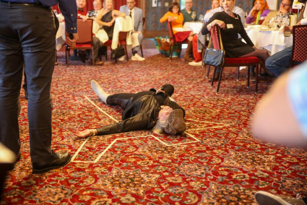 Actor lying on the floor as the “victim” in the middle of a Dinner Detective mystery scene while guests look on.