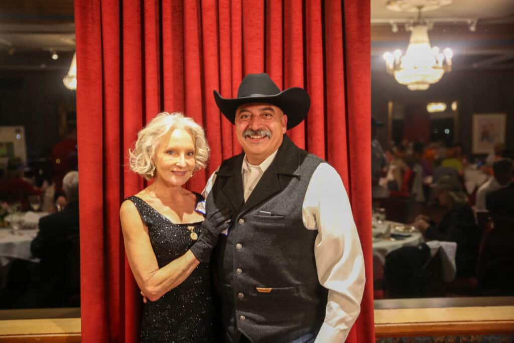 Smiling couple dressed up for a Dinner Detective mystery show, posing in front of a red theater curtain.
