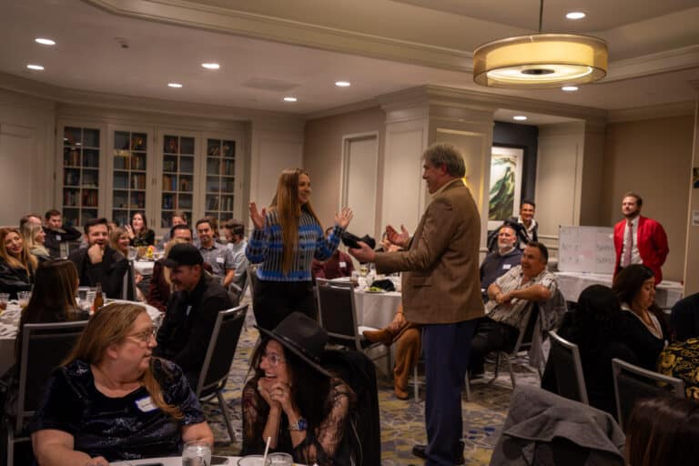 Corporate team members laughing and engaging during an interactive murder mystery dinner show with actors performing among seated guests at round tables in an upscale event venue