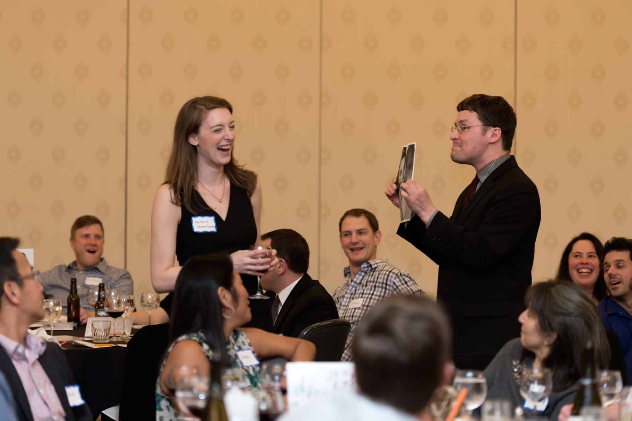 Guests laugh and interact with a performer during a Dinner Detective murder mystery show at a holiday event.
