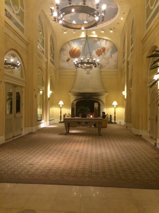 Elegant hotel lobby with high arched ceilings, large chandeliers, a painted sky and hot air balloon mural, a patterned carpet, and a central table with lamps and seating near a grand fireplace.