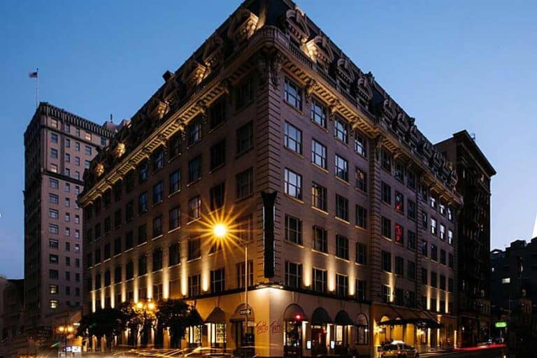 A large, historic hotel building on a city corner is illuminated by warm exterior lights at dusk. The sky is clear, and a few cars and streetlights are visible along the quiet streets.