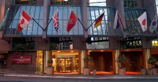 The entrance of a Crowne Plaza hotel, decorated with seven national flags above the doorway, with potted plants on either side and glass windows reflecting city buildings.