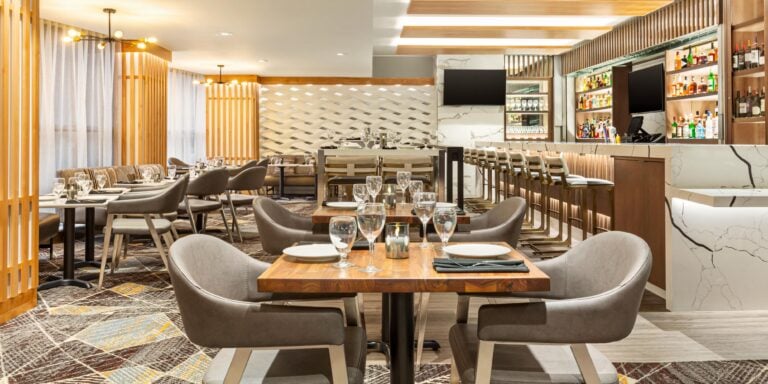 Modern restaurant interior with wooden tables, gray upholstered chairs, set dining tables with glassware, a bar area with stools, shelves stocked with bottles, and warm ambient lighting.