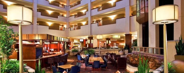 Spacious hotel lobby with modern lighting, indoor plants, a central seating area with tables and chairs, a bar on the left, and multiple floors with balcony railings overlooking the open atrium.