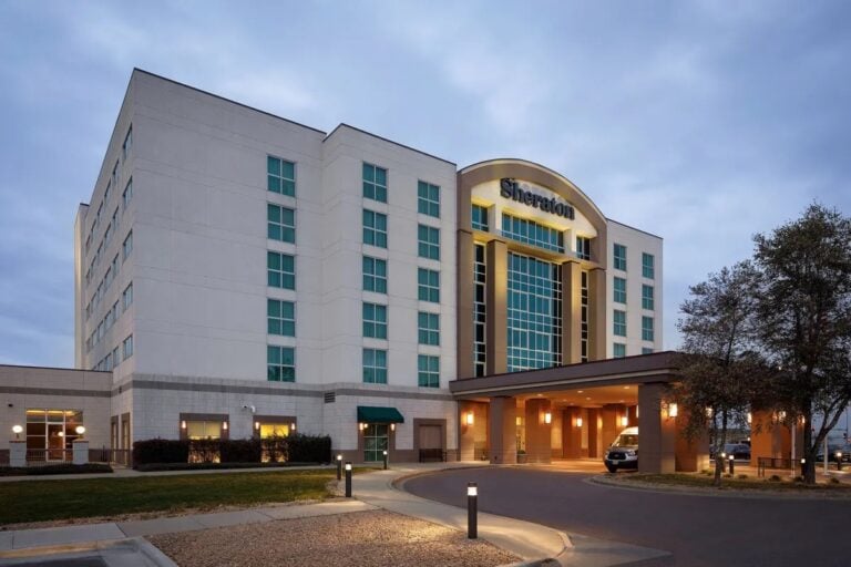 A modern, multi-story Sheraton hotel with large windows and a covered entrance, illuminated in the evening, with a car parked at the front and trees and a lawn nearby.