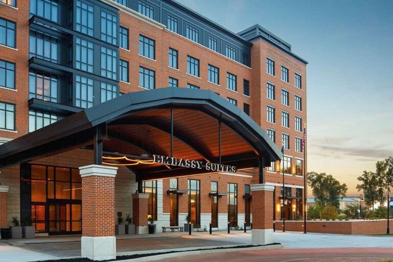 Front view of a modern hotel building with a red brick and black facade. The entrance features a large, arched canopy with glass doors, and the name Embassy Suites is visible. Trees and a clear sky are in the background.