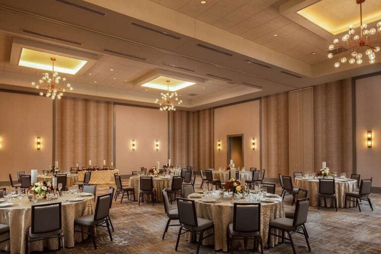A spacious banquet hall with round tables set for a formal event. Each table is adorned with floral centerpieces and table settings. Elegant chandeliers illuminate the room, and soft wall lighting enhances the warm, inviting atmosphere.