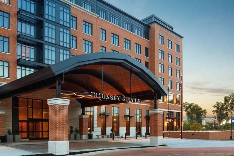 A modern, multi-story hotel with a brick and glass exterior, featuring a large covered entrance labeled Embassy Suites. The sky is clear and trees are in the background.