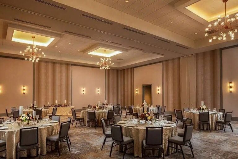 A banquet hall with round tables covered in gold tablecloths, set with plates and glasses, and decorated with floral centerpieces; elegant lighting fixtures hang from the ceiling.