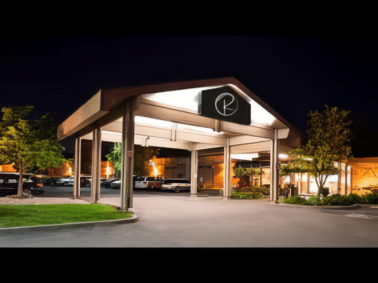 A modern building with a peaked roof, illuminated at night. The entrance features a circular logo with a stylized R. The surrounding area includes trees and a well-lit parking lot with cars.