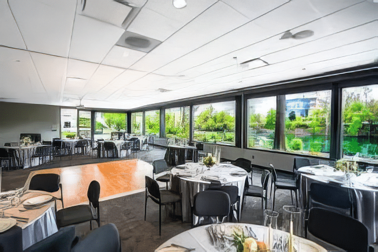 A bright banquet room with large windows offering a garden view. Several round tables are set with white tablecloths, plates, and cutlery. Black chairs surround the tables, and the floor is a mix of carpet and wooden dance area.