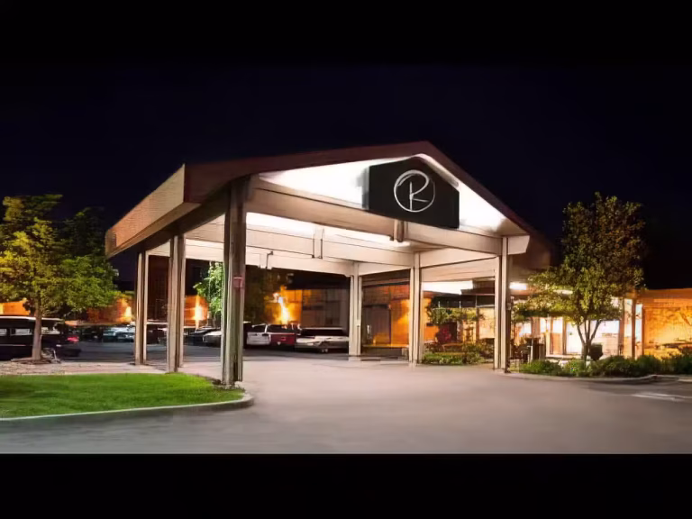 A well-lit building entrance with a peaked roof at night, featuring a circular logo above the entrance, surrounded by trees and a parking lot with visible parked cars.