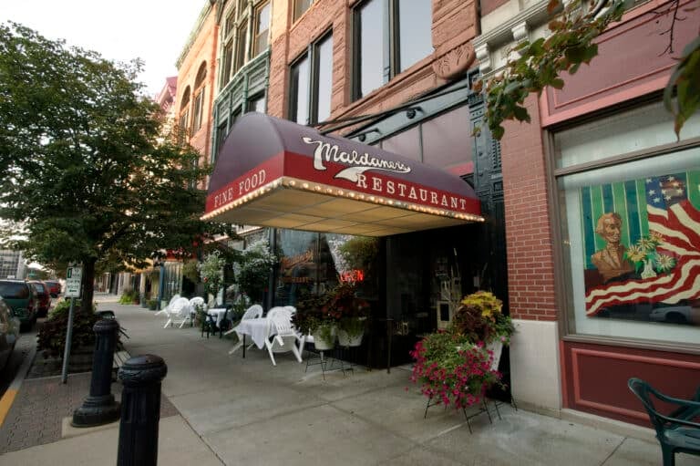 Street view of a restaurant with a red awning reading Maldaners Restaurant Fine Food. Outdoor seating with white chairs and tables is visible. A tree and building facade are in the background. A Wines sign is partially seen on a window.