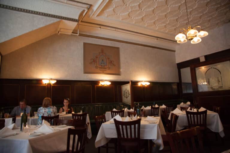 A dimly lit restaurant with vintage décor, featuring dark wood paneling and ornate ceiling details. Tables are set with white tablecloths and wooden chairs. Two people are seated at a table on the left. A chandelier hangs overhead.