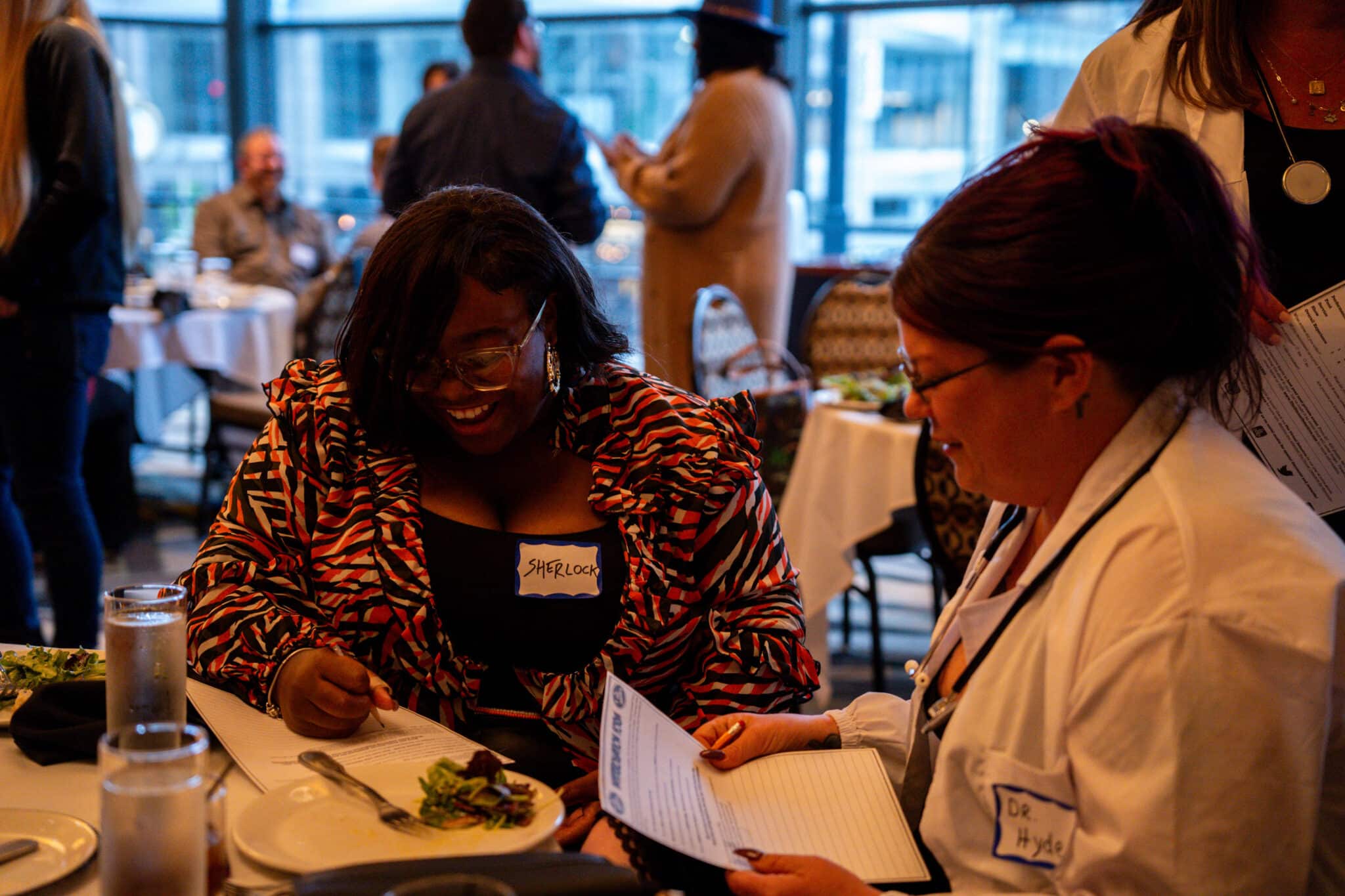 Two women sit at a table in a lively restaurant, smiling and writing on papers. One wears a patterned jacket and glasses, the other a white coat; both have name tags. Other people and tables are visible in the background.