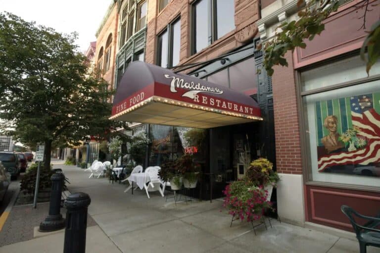 A view of Maldaners Restaurant with a red awning, outdoor tables set with white chairs, potted plants, and a colorful mural of a man and an American flag on the building’s brick facade.