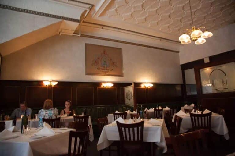 A dimly lit restaurant dining area with white tablecloths, wooden chairs, wall lamps, and a decorative ceiling. Three people sit at one table, while the other tables are empty. A framed artwork is on the wall.