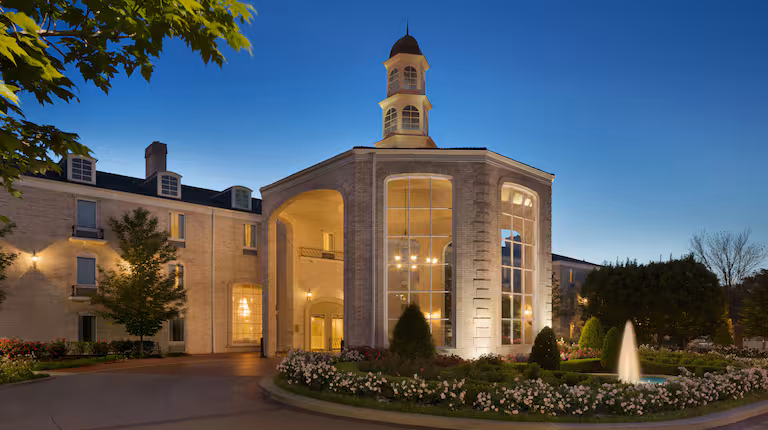 A grand hotel entrance at dusk, featuring tall arched windows, a small cupola tower, and a circular driveway with manicured gardens and a fountain in the foreground, all warmly lit.