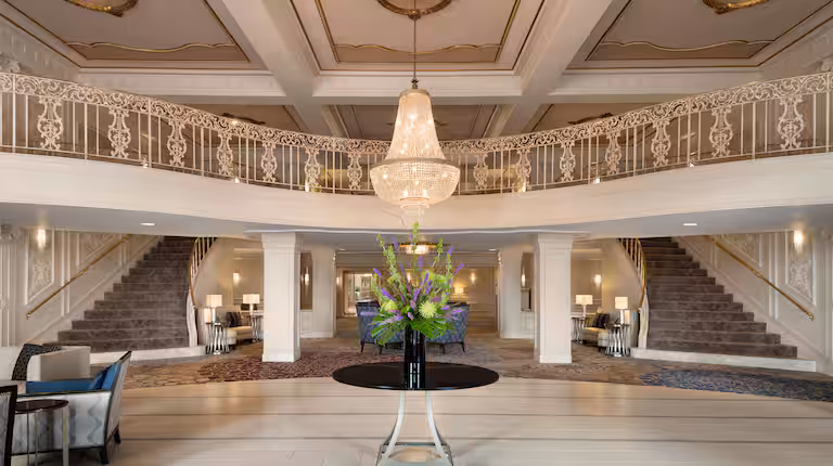 Grand hotel lobby with a large chandelier, elegant staircase splitting to both sides, ornate railings, and a round table with a floral arrangement in the center. The decor is classic and sophisticated.