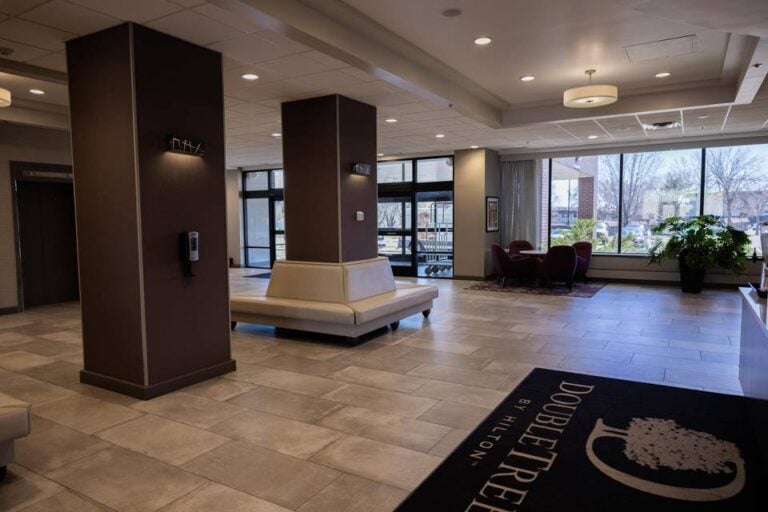 A spacious hotel lobby with tiled floors, modern seating, large windows letting in natural light, potted plants, and a black rug with the DoubleTree by Hilton logo near the entrance.
