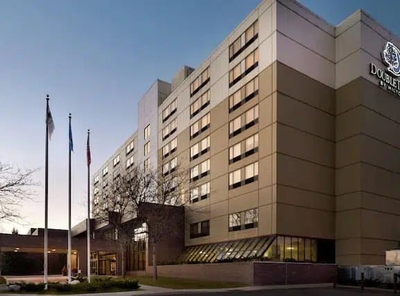 A modern multi-story hotel building with large windows at dusk, featuring the DoubleTree by Hilton logo. Several flagpoles with flags are in front, and leafless trees are visible near the entrance.