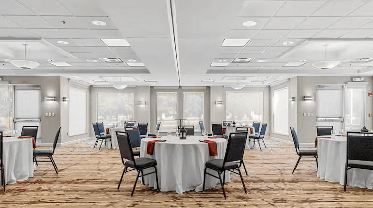 A banquet hall with round tables draped in white tablecloths and orange accents is set for an event, as if preparing for a true crime mystery show. Several chairs encircle each table in a well-lit room with large windows, adding a touch of intrigue.