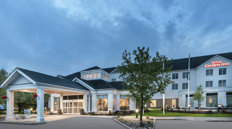 The exterior view of a Hilton Garden Inn hotel, with its gray roof and white facade, hints at a true crime mystery unfolding under the cloudy sky. The entrance canopy and illuminated sign are surrounded by trees, casting shadows on the paved driveway below.