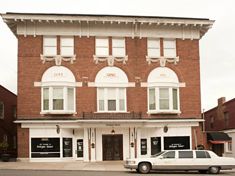 A historic red-brick building with ornate white trim reading 1845 and 1911 above arched windows. A white stretch limousine is parked in front. The ground floor has signs for Michigan Manor.