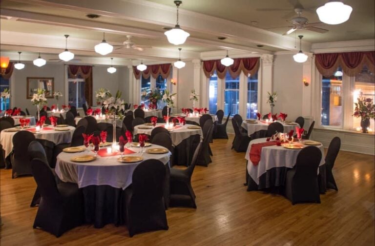 Elegant banquet hall with round tables covered in white and black cloths, red napkins, gold plates, lit candles, and floral centerpieces. Large windows with red curtains and wooden floors complete the setting.