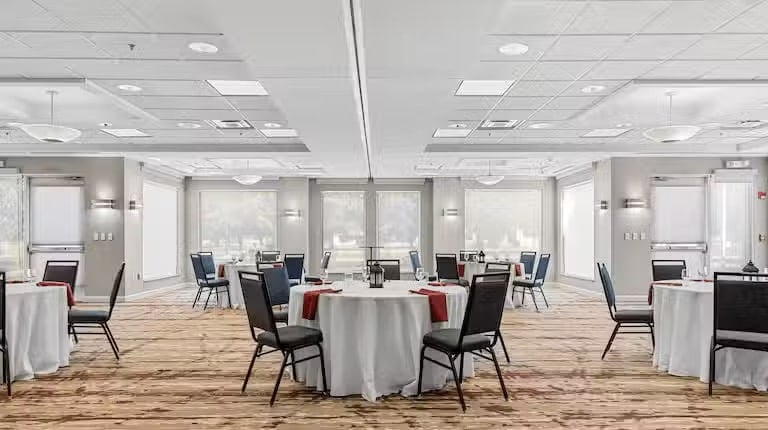 A spacious, well-lit conference room with round tables covered in white tablecloths and surrounded by chairs. Each table has a red napkin and a centerpiece. Large windows let in natural light.
