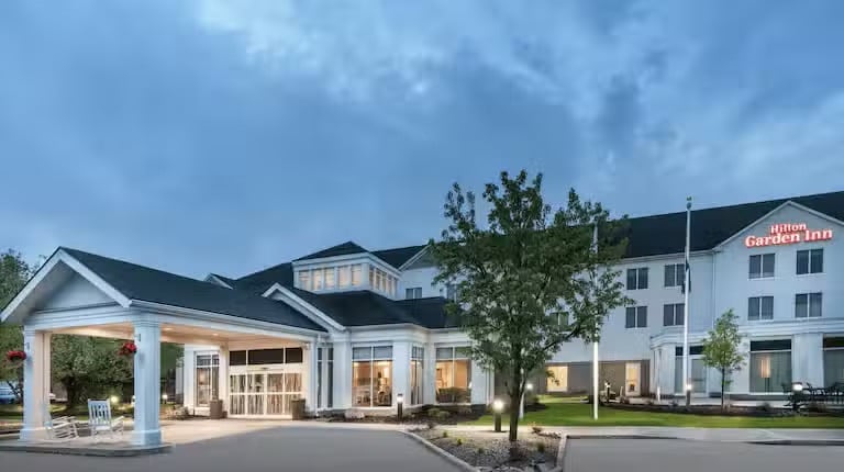 Exterior view of a Hilton Garden Inn hotel at dusk, with illuminated windows, a covered entrance, and a few trees in the landscaped front area, set against a partly cloudy sky.